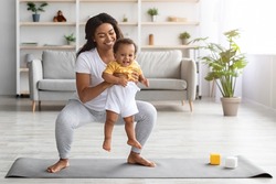 Weight Loss After Pregnancy. Happy Black Mom Training At Home With Infant Baby, Making Squats While Holding Her Cute Toddler Son In Hands, Exercising Pilates On Fitness Mat In Living Room, Copy Space