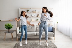 Playful korean mother and pretty daughter dancing to music and jumping at home. Asian mom and her kid enjoying favorite song together, fooling around, having fun in living room interior