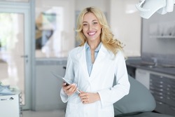 Young beautiful woman in workwear dentist holding brand new digital tablet, cheerfully smiling at camera, doc using modern technologies while planning treatment, online appointment with dental clinic