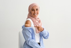 Portrait Of Vaccinated Muslim Woman In Hijab Pointing On Arm With Adhesive Bandage After Coronavirus Vaccine Injection Shot, Smiling Islamic Lady Posing On White Studio Background, Copy Space
