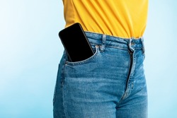 Unrecognizable woman having smartphone with empty screen in her jeans pocket on blue studio background, mockup for mobile app or website design. Space for your ad template