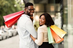 Sale And Consumerism Concept. Portrait of happy African American couple holding shopping bags. Smiling black man and woman carrying purchases, turning back and looking at camera over the shoulder