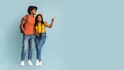 Happy millennial black couple tourists with backpacks and camera embracing and looking at copy space on blue studio background, choosing destination to travel, panorama. Travelling concept