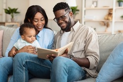Early Development for Babies. Portrait of millennial black parents reading book to their adorable toddler baby boy at home, sitting on couch in cozy living room, spending time together