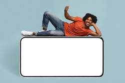 Cheerful african american young man relaxing on huge horizontal brand new cellphone with empty screen and showing thumb up, mockup, black guy enjoying newest entertaining mobile app