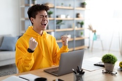 Excited asian man emotionally celebrating success using laptop in home office, sitting at desk raising hands with joy, shaking fists, received good news, got promotion, finished project, free space