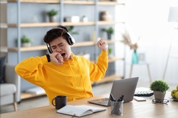 Tired asian male student yawning sitting at desk using laptop. Bored youth is exhausted from getting ready for test or writing coursework, feeling sleepy, stretching wearing headphones. Lack of sleep