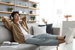 Rest And Relax Concept. Calm asian man sitting on couch, listening to music, audio book, podcast, enjoying meditation for sleep and peaceful mind in wireless headphones, leaning back, copy space