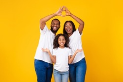 Love, Care And Support. Smiling black parents showing heart shape with fingers, connecting their hands together, making symbolic roof under their daughter head isolated on yellow orange studio wall