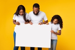 Cheerful African American man, woman and girl looking down at empty advertising placard, yellow studio background. Smiling black family holding blank board for advertisement or text in hands