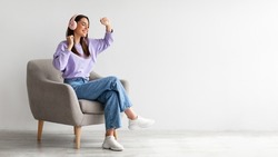 Cheery young lady in headphones listeing to music and dancing while sitting in armchair against white studio wall, banner design with free space. Carefree Caucasian woman moving to favorite song