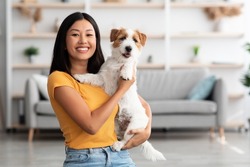 Portrait of cheerful young asian woman in casual outfit carrying her sweet fluffy dog jack russel terrier breed, pet and owner having good time together at home, living room interior, copy space