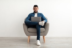 Young Arab man in casual wear working online, sitting in armchair and using laptop against white studio wall, full length. Millennial Eastern guy surfing internet on portable pc