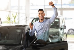 Emotional handsome middle-eastern guy raising hand up and smiling at camera, showing key from his brand new black car, copy space. Rich arabic man buying auto at luxury dealership salon