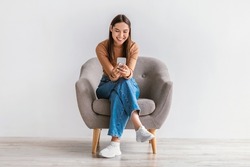 Happy Caucasian woman using cellphone, chatting on internet, working or studying online, sitting in armchair against white studio wall, full length. Cheery young lady watching video on mobile phone