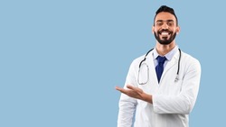 Happy Arabic Doctor Man Smiling Showing Copy Space With Hand Advertising Your Text Over Blue Studio Background. Portrait Of Successful Physician. Medical Advertisement Banner