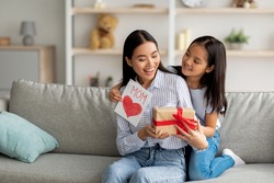 Congratulations. Loving asian daughter greeting surprised mom with Mother's Day, giving her handmade card and gift box, sitting on sofa at home interior, free space