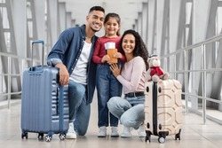 Happy Middle Eastern Family Of Three Posing In Airport Terminal, Cheerful Arab Parents And Their Cute Little Daughter Holding Suitcases, Passports And Tickets And Smiling At Camera, Ready For Travel
