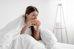 Starving upset hungry cute caucasian millennial woman sitting in bed at home and eating burger suffering from stress, depression, crisis and gluttony. Junk food, substitution, diet and too much meal