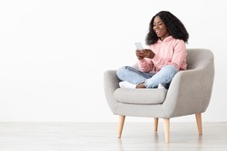 Cheerful african american young lady sitting in arm chair at home, using nice mobile application on brand new smartphone, shopping online, white background, panorama with copy space
