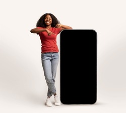 Cheerful Young African American Lady Leaning And Pointing At Big Smartphone With Blank Screen, Smiling Black Lady Showing Copy Space For Advertisement, Standing Over White Studio Background, Mockup