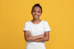 Happy calm teen black girl pupil in white t-shirt with crossed arms, isolated on yellow background, empty space. Positive schoolgirl, student emotions, good ad and covid-19 quarantine, studio shot