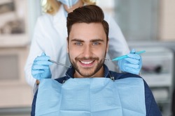 Smiling young bearded man sitting in dental chair, visiting modern dental clinic, having regular checkup, unrecognizable woman dentist holding dental tools next to male patient mouth