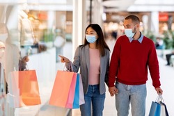 Young diverse couple in face masks shopping at supermarket, holding gift bags, pointing at shop window, selecting goods, copy space. Millennial family staying safe at mall during coronavirus pandemic