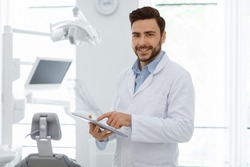 Attractive male dentist cheerful young bearded man with newest digital tablet smiling at camera, doctor using modern technologies while planning treatment, online appointment with dental clinic