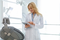Young blonde woman dentist standing by dental chair, using digital tablet at newest modern dental clinic, chatting with client or checking her schedule online, copy space. Medical appointment concept
