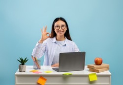 Happy asian female student in eyeglasses showing okay sign gesture sitting at the desk and using laptop over blue background. Lady doing homework, recommending online school or educational course