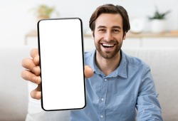 Excited Young Man Demonstrating Smartphone With Big Blank White Screen At Camera, Handsome Smiling Millennial Guy Showing Copy Space For Mobile App Advertisement, Sitting On Couch At Home, Mockup