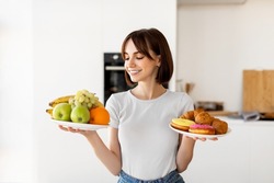 Choosing between healthy or unhealthy food. Young woman holding plates with fruits and sweets, looking at healthy snack with smile, deciding what to eat, standing in kitchen interior