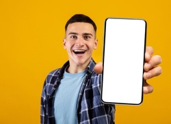 Cheerful Young Guy Demonstrating Smartphone With Big Blank White Screen, Excited Man Showing Free Copy Space For Your Ad, Recommending New Mobile App, Standing Over Yellow Background, Mockup