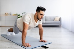 Strength workout concept. Fit young Arab man doing push ups or plank at home, following video tutorial on smartphone, free space. Athletic Eastern guy having domestic training during covid-19 lockdown