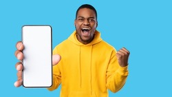 Emotional african american guy gamer showing smartphone with blank screen and celebrating success, trading online, using mobile application, blue studio background, panorama, copy space, mockup