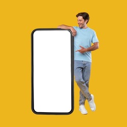 Mobile App Advertisement. Full Body Length Of Happy Man Leaning And Pointing At Big Huge White Empty Smartphone Screen Standing On Orange Studio Background. Check This Out, Cellphone Display Mock Up