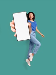 Full length of cheery Asian woman jumping and smiling, showing cellphone with empty space for mobile app or website on screen, turquoise studio background, mockup. Creative collage