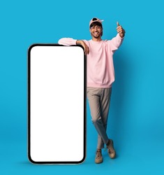 Positive stylish indian guy posing with big smartphone with empty screen, showing thumb up and smiling, recommending newest mobile app, blue studio background, mockup, full length shot