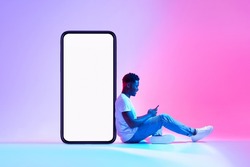 Young black man sitting next to huge smartphone with empty screen, advertising new mobile application or website, using modern gadget in neon light. Cellphone display mockup