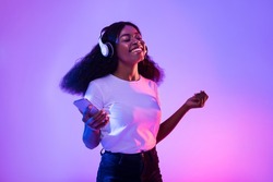 Happy African American woman with headphones and smartphone listening to music and dancing in neon light. Young black woman moving to favorite song, enjoying cool playlist