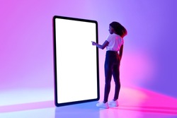 Millennial African American woman touching screen of huge tablet computer, interacting with pc interface in neon light, mockup for new app or website design. Touch pad template