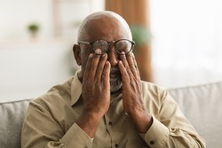 Glaucoma. Senior African American Man Rubbing Tired Eyes Wearing Eyeglasses Having Poor Eyesight Sitting On Couch At Home. Ophtalmology Diseases, Health Problems In Older Age Concept