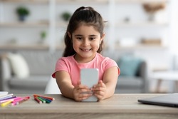Gadgets And Kids. Cute Little Arab Girl Using Smartphone At Home, Adorable Female Child Playing Mobile Games Or Watching Cartoons Online On Cellphone While Sitting At Table In Living Room, Closeup