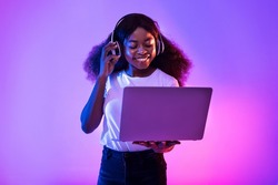 Young black woman having video call, using laptop computer, wearing wireless headphones in neon light. Happy African American lady watching movie, chatting online on pc