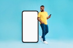 Cheery millennial black guy with mobile device looking at giant smartphone on blue background, mockup for mobile app on white screen. Cellphone website template, cool online store ad
