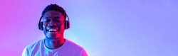 Handsome young black man wearing headphones, listening to music with closed eyes in neon light, banner design. Happy African American guy enjoying new playlist and smiling