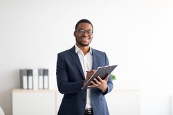 Happy young black male psychologist writing in clipboard, looking at camera and smiling in modern office. Psychotherapy services, mental health professional concept