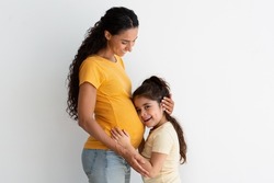 Happy Expectation. Adorable Little Preteen Girl Cuddling Mother's Pregnant Belly, Excited Curious Cute Female Child Awaiting For Sibling, Hugging Mommy's Tummy While Bonding Together At Home