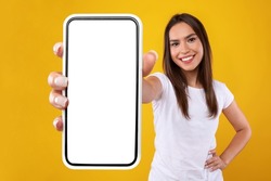 App Ad. Smiling pretty woman holding smartphone with white blank device screen in hand close up to camera, orange studio. Gadget with empty free space for mockup, banner isolated, selective focus